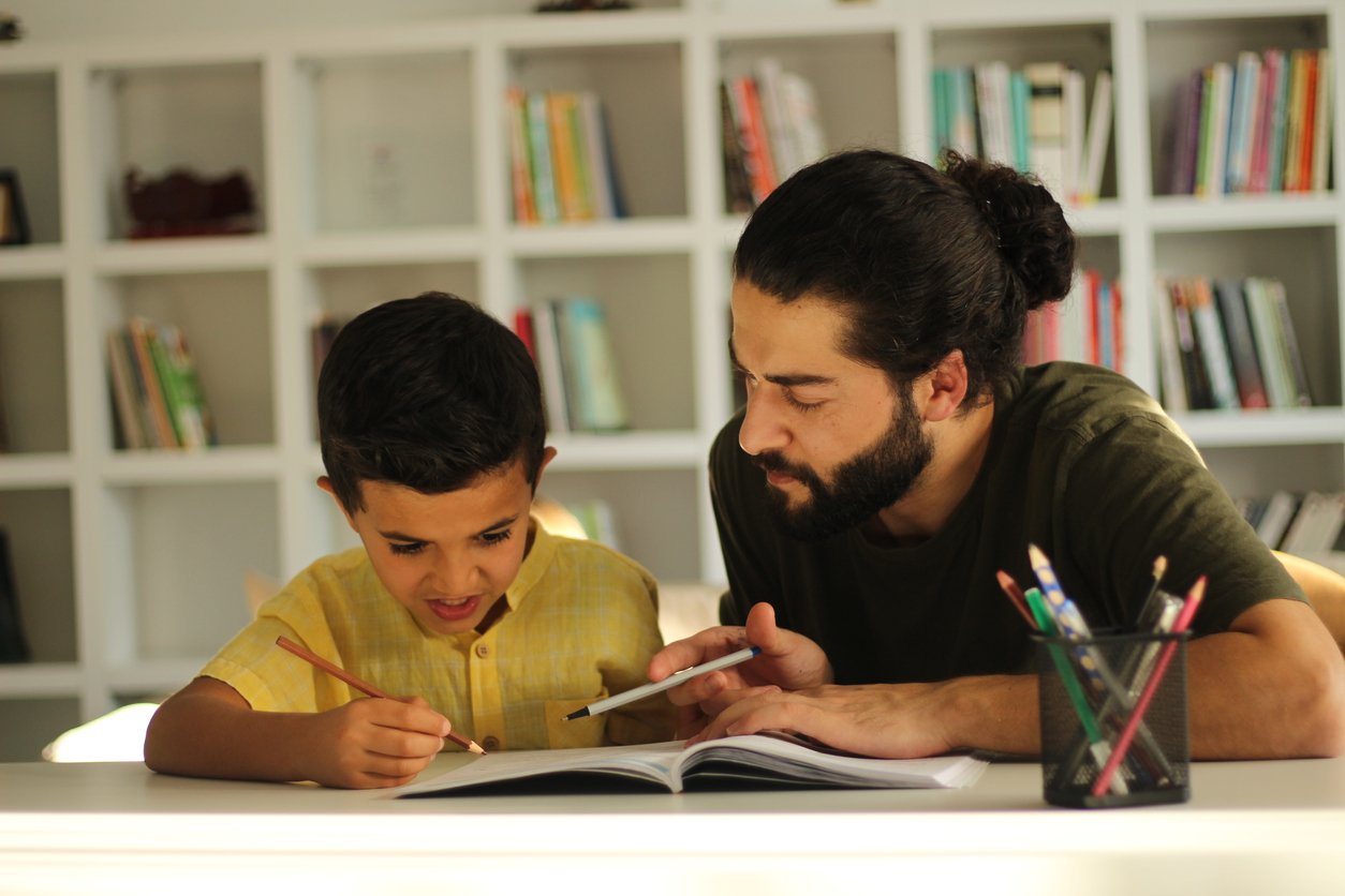 Look here. Happy delighted good looking man sitting with his son and help him for doing homework